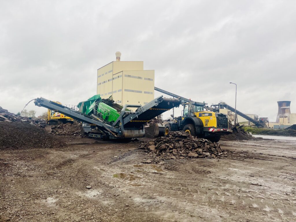 JCB excavator levelling soil on a construction site with a large spoil area and cloudy sky in the background.