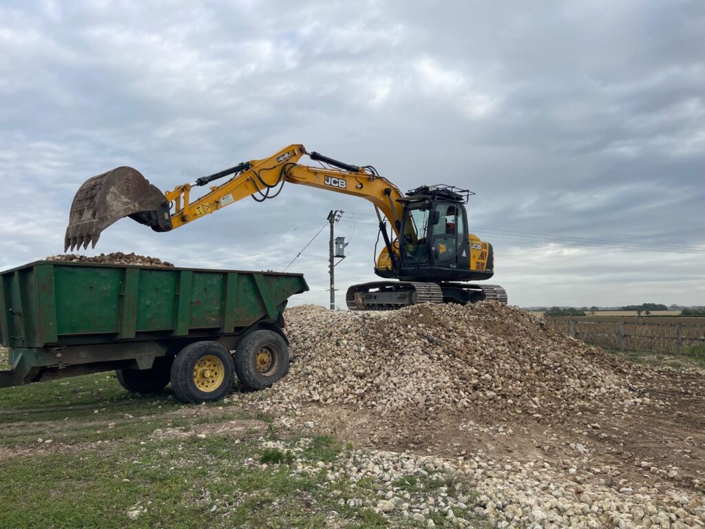 Tracked JCB excavator loading stone into a green dump trailer on a rural site.