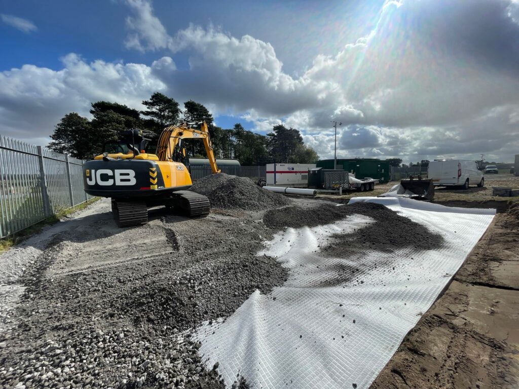 Tracked JCB excavator spreading aggregate across a geotextile base layer.
